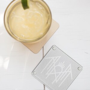a glass of yellow liquid with a lime wedge, placed on a white wooden surface next to a coaster with a graphic design.