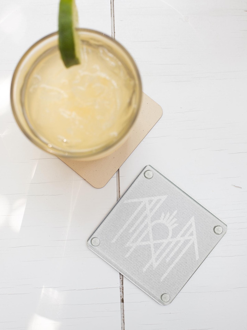 a glass of yellow liquid with a lime wedge, placed on a white wooden surface next to a coaster with a graphic design.