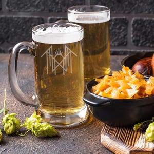 two glasses of beer, one with a logo on it, placed on a wooden surface alongside a bowl of food and some hop cones.