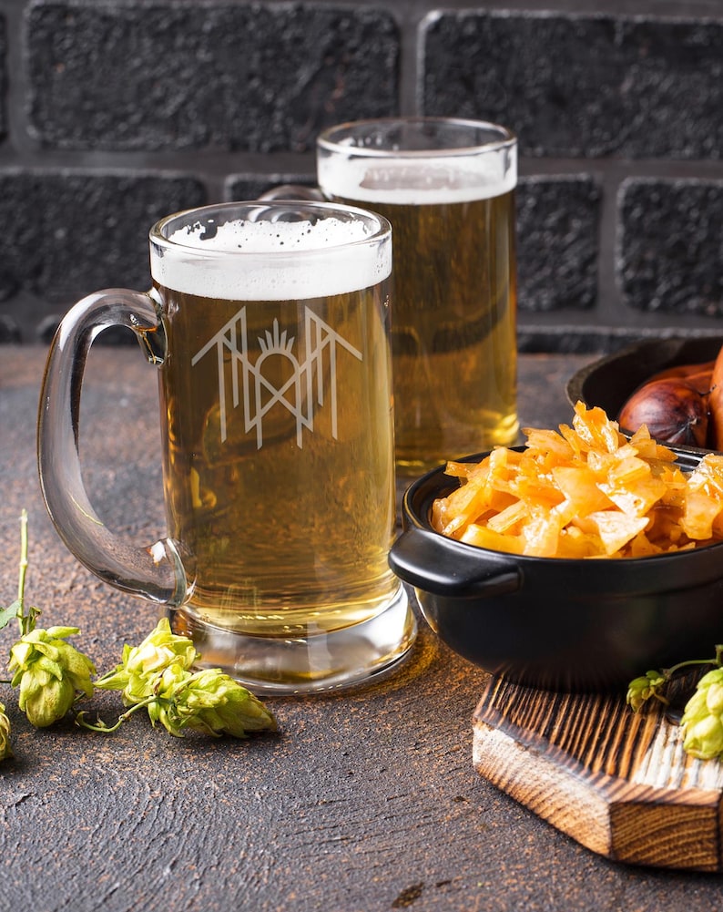 two glasses of beer, one with a logo on it, placed on a wooden surface alongside a bowl of food and some hop cones.