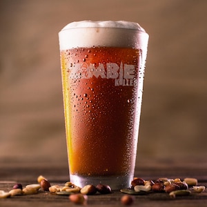 May include: A pint glass filled with amber beer, topped with a creamy white head. The glass is etched with the words "ZOMBIE KULLER" and is covered in condensation. Mixed nuts are scattered on the wooden surface in the foreground.