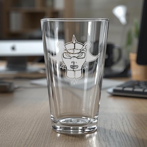 May include: A clear pint glass featuring a white etched design of a stylized face with a helmet. The glass is empty and stands on a wooden desk. The background includes a computer monitor, keyboard, and other office items.