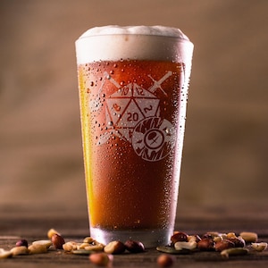 May include: A pint glass filled with amber beer and a foamy head. The glass is etched with a design featuring a 20-sided die, swords, and a shield. The glass has condensation, and mixed nuts are scattered at the base. The glass is approximately 15 cm tall.