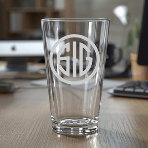 May include: A clear pint glass with a white monogram design. The monogram features the letters S, I, and G within a circular border. The glass is empty and sits on a wooden desk.