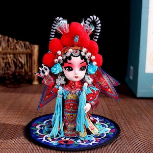 Könnte beinhalten: Eine dekorative Puppe in einem traditionellen rot-blauen Opernkostüm. Die Puppe trägt einen Kopfschmuck mit großen roten Pompons, weißen Perlen sowie schwarz-weiß gestreiften Akzenten. Sie trägt ein rotes, gemustertes Gewand mit langen blauen Quasten.
