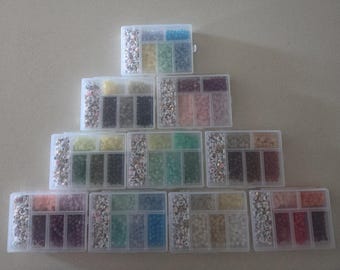 Può includere: Dieci contenitori in plastica trasparente disposti a piramide, ciascuno suddiviso in più scomparti riempiti con un assortimento di piccole perline colorate, tra cui perline rotonde a tinta unita e perline bianche con lettere nere stampate.