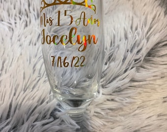 May include: A clear champagne flute with gold lettering and a gold crown design. The text on the glass reads "Mis 15 Años Jocelyn" and "7/16/22". The glass is designed for a quinceañera celebration.