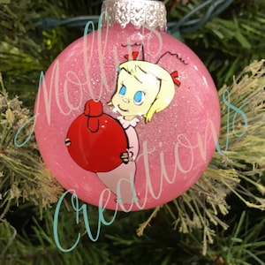 May include: Pink glitter Christmas ornament featuring a cartoon character holding a red ornament. The ornament has a silver metal cap and a loop for hanging. The text "Molly's Creations" is written on the ornament.