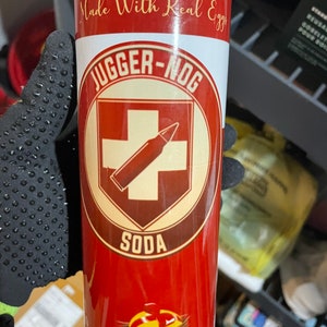 May include: A red and white insulated tumbler with a white circle containing a red cross and a brown bullet. The text "JUGGER-NOG SODA" is printed on the tumbler. The text "Made With Real Eggs" is printed on the top of the tumbler. The text "12 FL. OZ (355ML)" is printed on the bottom of the tumbler.
