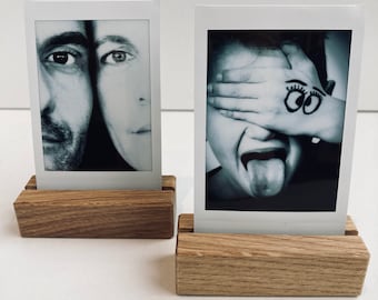 Könnte beinhalten: Zwei Schwarzweiß-Polaroidfotos werden in Holzrahmen angezeigt. Die Fotos zeigen zwei verschiedene Personen. Das erste Foto zeigt das Gesicht eines Mannes und einer Frau nebeneinander. Das zweite Foto zeigt eine Person, die sich die Augen mit der Hand bedeckt und die Zunge herausstreckt. Das Foto hat eine Zeichnung von zwei Augen auf der Hand.