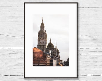 Puede incluir: Una fotografía enmarcada de una ciudad con un alto campanario de iglesia puntiagudo en el centro. La iglesia está hecha de piedra y tiene una cruz en la parte superior. Los edificios en el fondo también están hechos de piedra y tienen un diseño más moderno.