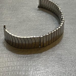 May include: A silver metal watch band with a flexible design. The band is made of small, interlocking metal links.