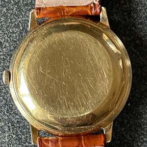 以下が含まれることがあります： 茶色の革バンドが付いたゴールドトーンの腕時計。時計の文字盤は透明なガラスで覆われています。