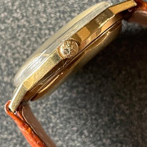 以下が含まれることがあります： 茶色の革バンドが付いたゴールドトーンの腕時計。時計は丸い文字盤と側面に小さなリューズがあります。