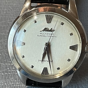 Puede incluir: Un reloj Mido Multifort Superautomatic plateado con una esfera blanca y una correa de cuero negra. La esfera del reloj tiene marcadores de hora en números romanos negros y una aguja de segundos negra. La esfera del reloj también tiene el texto "Mido Multifort Superautomatic" y "powerwind".
