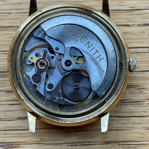 以下が含まれることがあります： 金色調の腕時計の裏側。銀色の文字盤には「ZENITH」と「SWISS MADE」の文字が印刷されています。この時計は30石のムーブメントを搭載しています。