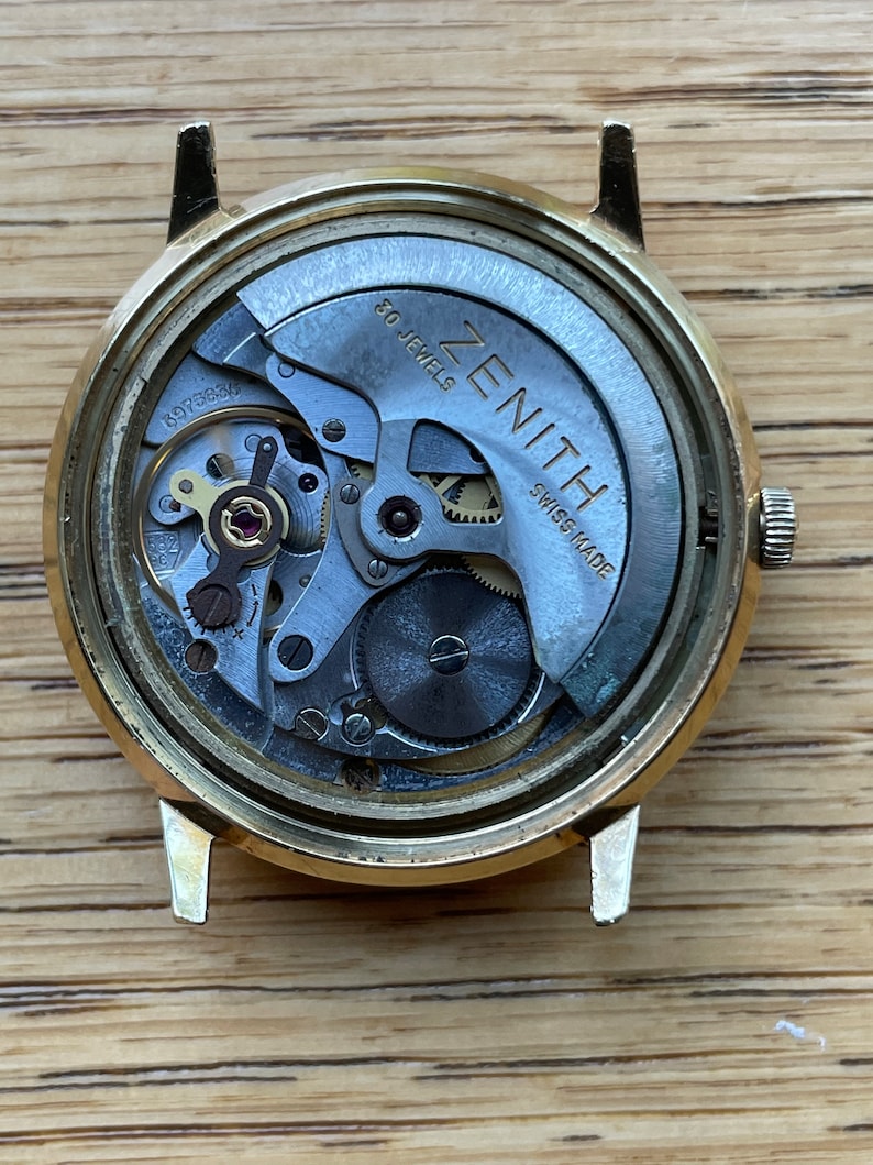以下が含まれることがあります： 金色調の腕時計の裏側。銀色の文字盤には「ZENITH」と「SWISS MADE」の文字が印刷されています。この時計は30石のムーブメントを搭載しています。
