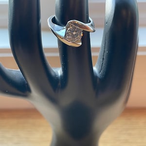 Könnte beinhalten: Ein silberner Ring mit einem großen, klaren, runden Edelstein, der in einem verdrehten Band gefasst ist.