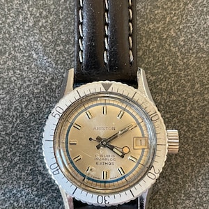 Puede incluir: Un reloj de pulsera plateado con una correa de cuero negro. La esfera del reloj es blanca con marcas negras y las palabras "ARISTON" y "17 RUBIS INCABLOC 5 ATMOS" impresas en ella.