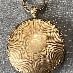 Może przedstawiać: Złoty medalion z okrągłym, fakturowanym wzorem i ozdobną ramką. Medalion ma pierścień u góry do przymocowania łańcuszka.