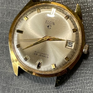 Op de afbeelding: Een gouden Elgin automatische polshorloge met een witte wijzerplaat en zwarte wijzers. De wijzerplaat van het horloge heeft een datumvenster op 3 uur en de tekst 'ELGIN E' en 'AUTOMATIC 25 JEWELS'.