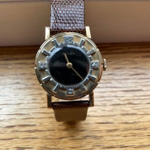 May include: A gold-toned wristwatch with a black face and diamond-shaped hour markers. The watch has a brown leather band with a textured pattern.