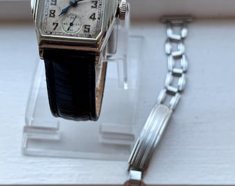 Può includere: Un orologio da polso Waltham rettangolare vintage con quadrante color crema, cinturino in pelle nera e cassa argentata. L'orologio ha un piccolo quadrante dei secondi. Accanto c'è un cinturino in metallo argentato.