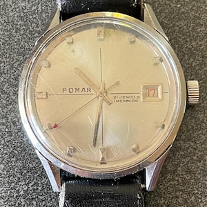 Puede incluir: Un reloj de pulsera Pomar vintage con esfera plateada, con el texto "POMAR" y "21 JEWELS INCABLOC". El reloj tiene una pantalla de fecha a las 3 en punto y está colocado en una correa de cuero negro.