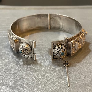 May include: A silver hinged bangle bracelet with intricate filigree designs and turquoise stones. The bracelet has a small chain with a key charm.