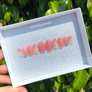 K&ouml;nnte beinhalten: Ein Set mit zehn pinkfarbenen und wei&szlig;en Ombre-Press-on-N&auml;geln mit goldenem Glitzer und Stern-Akzenten. Die N&auml;gel werden in einer wei&szlig;en Box mit dem Text "@pinkypresssed" auf der Vorderseite pr&auml;sentiert.