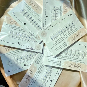May include: Sizing kit for press-on nails. The kit includes multiple clear plastic nail tips with numbers on them. The text on the card reads "Sizing Kit" and "Test each tip on your natural nail to find the best fit. The press-on should rest comfortably from edge to edge, without applying any pressure. Record your sizes below from THUMB to PINKY."