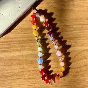 May include: A colorful beaded phone charm with a variety of gemstone beads in a rainbow of colors. The charm is attached to a silver laptop. The beads are round and irregular in shape, with gold-colored spacers.