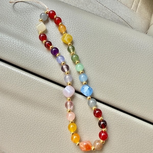 May include: A colorful beaded phone charm featuring a rainbow of gemstones. The charm includes red, orange, yellow, green, blue, and purple beads, with gold-colored spacers. The charm is attached to a thin, off-white cord.