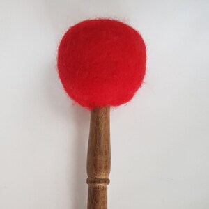 May include: A wooden mallet with a red, spherical head. The handle is a light brown color and has a rounded shape. The mallet is designed for striking instruments, such as singing bowls or gongs.