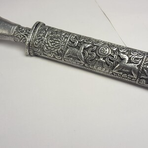 May include: A silver-colored, ornate dagger with detailed relief carvings. The handle and sheath are covered in intricate patterns, including floral designs and depictions of animals. The dagger is displayed on a white surface.