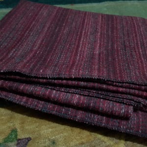 May include: Folded burgundy fabric with a striped pattern. The fabric has a textured appearance with dark and light threads woven together. The edges are finished with a dark, stitched border. The fabric is stacked on top of each other.
