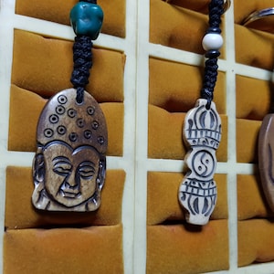 Op de afbeelding: Een verzameling gesneden houten hangers met zwarte koorden en metalen sleutelhangers. De hangers hebben verschillende ontwerpen, waaronder een Boeddha-gezicht, een yin-yang-symbool en een Om-symbool. De achtergrond is een display met een geel-oranje stof.