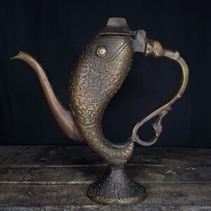 May include: An antique brass teapot with a fish-like design. The teapot has detailed floral patterns and a curved spout. The handle is also curved, and the base is a pedestal shape. The teapot is a dark brass colour.