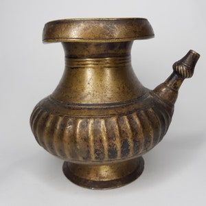 May include: An antique bronze-coloured pitcher with a bulbous body and a flared spout. The vessel has a wide, flat rim and a decorative, ribbed pattern around the midsection. The spout is adorned with a twisted rope-like detail.