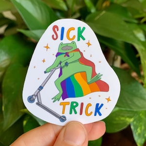 May include: A white sticker with a green frog wearing a rainbow cape and riding a scooter. The sticker says "SICK TRICK" in rainbow letters.