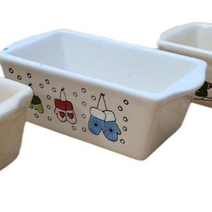 May include: Three ceramic loaf pans with different designs. The first pan has a green olive branch design. The second pan has a winter-themed design with mittens and snowflakes. The third pan has a snowman design.