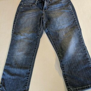 May include: Dark wash denim jeans with a classic five-pocket design. The jeans have a straight leg cut and a button and zipper closure. The denim has a faded appearance, with lighter areas on the thighs and knees. The jeans are laid flat on a light-colored surface.