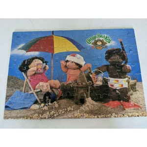 May include: A vintage Cabbage Patch Kids puzzle featuring three dolls on a beach scene. The dolls are wearing summer clothes and are playing in the sand. The puzzle is made of cardboard and has a blue background with a colorful umbrella.