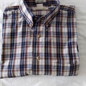 May include: A folded button-down shirt with a plaid pattern in navy blue, burgundy, and white. The shirt has a button-down collar and a pocket on the left side. The tag on the collar reads "Wrinkle Free".