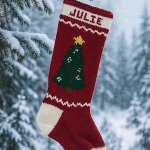 Peut inclure: Une chaussette de Noël rouge et blanche tricotée avec le nom "JULIE" en haut. La chaussette présente un motif de sapin de Noël vert avec une étoile jaune et des accents blancs. Elle est suspendue à une branche de pin enneigée.