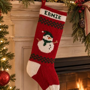 May include: A red knitted Christmas stocking featuring a white cuff with the name ERNIE embroidered in green. The stocking displays a central snowman graphic wearing a green hat and scarf, accented by green checkered patterns above and below.