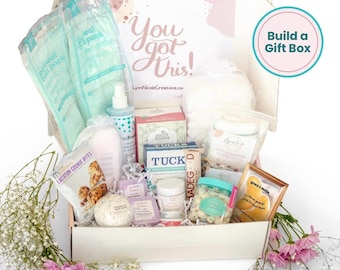 May include: A white cardboard gift box filled with various wellness products including cold packs, lactation cookie bites, a spray bottle, Tucks pads, a small jar, and skincare items. The box lid features the text You got this! and Build a Gift Box.