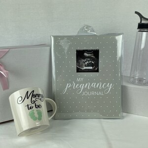 May include: A gift set for a new parent includes a white mug with the text "Mommy to be" and green footprints, a gray journal with the text "My Pregnancy Journal" and a clear water bottle with a black lid.