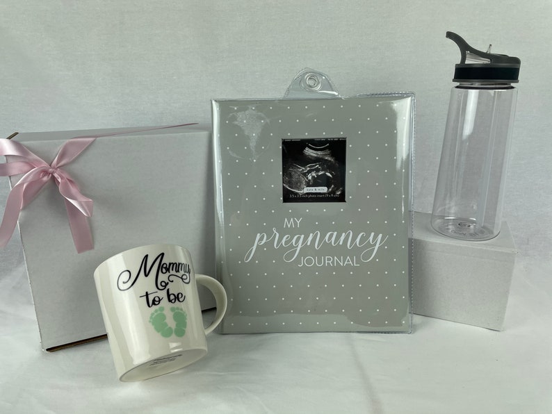 May include: A gift set for a new parent includes a white mug with the text "Mommy to be" and green footprints, a gray journal with the text "My Pregnancy Journal" and a clear water bottle with a black lid.