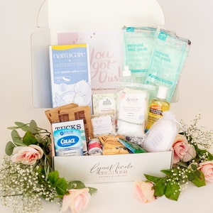 May include: A white gift box filled with various postpartum care items, including perineal cold packs, maternity pads, a stool softener, and a bottle of cocoa butter. The box is decorated with pink roses and the text "Lynn Nicole Creations New Mom & Self Care Gifts".